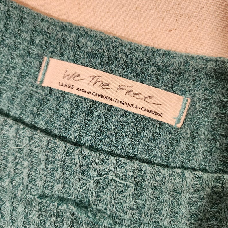 Puede incluir: Primer plano de una tela de punto de gofre de color verde azulado. Una etiqueta blanca con el texto "We The Free" est&aacute; cosida a la tela. La etiqueta tambi&eacute;n incluye la talla "LARGE" y el texto "MADE IN CAMBODIA / FABRIQU&Eacute; AU CAMBODGE".