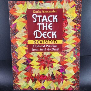 May include: A book titled "Stack the Deck Revisited" by Karla Alexander. The cover features a colorful patchwork design with red, yellow, and orange hues. The book's title is in large, white, stylized letters on a burgundy background.