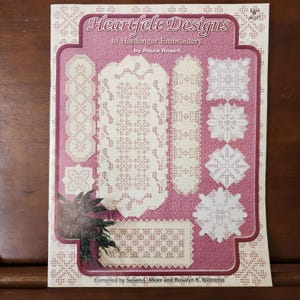 May include: A book titled "Heartfelt Designs in Hardanger Embroidery" by Paula Roach. The cover features a pink background with white embroidered designs, including a table runner, placemats, and napkins.