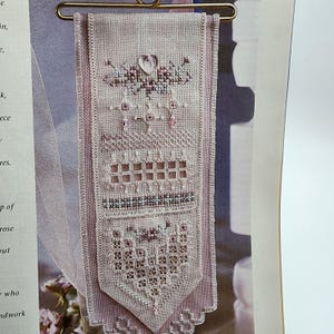 May include: A detailed, hand-stitched cross-stitch wall hanging in shades of pink and white. The design features floral and geometric patterns, including hearts and squares. The piece is displayed on a gold-colored hanger.