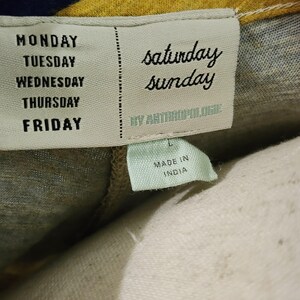 Puede incluir: Primer plano de una etiqueta de ropa con los d&iacute;as de la semana impresos verticalmente en el lado izquierdo. Las palabras "saturday sunday" est&aacute;n impresas en cursiva en el lado derecho. La etiqueta tambi&eacute;n dice "BY ANTHROPOLOGIE" y "MADE IN INDIA".