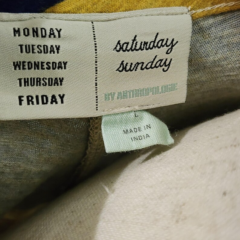 Puede incluir: Primer plano de una etiqueta de ropa con los d&iacute;as de la semana impresos verticalmente en el lado izquierdo. Las palabras "saturday sunday" est&aacute;n impresas en cursiva en el lado derecho. La etiqueta tambi&eacute;n dice "BY ANTHROPOLOGIE" y "MADE IN INDIA".