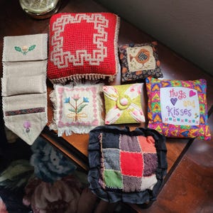 May include: A collection of handmade decorative pillows in various shapes and sizes. Pillows feature cross-stitch and patchwork designs, including floral patterns, geometric borders, and the text "hugs and kisses". Colors include red, green, yellow, and blue.