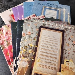 May include: A collection of vintage needlework books and framed samplers. The books have floral and patterned covers, with titles like "Heirloom Drawn Thread Sampler" and "Miniature Drawn Thread Sampler." The framed sampler displays intricate stitch patterns.