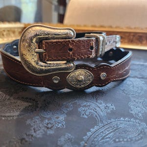 May include: A brown leather belt with a silver buckle and a decorative silver oval. The belt has a scalloped edge and rhinestones.