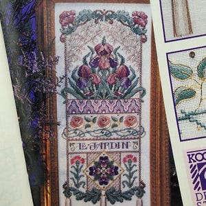 May include: Cross-stitch floral design featuring purple irises, roses, and other flowers, framed in a wooden border. The design includes the text "Le Jardin". The needlework is detailed and colorful, showcasing a garden theme.