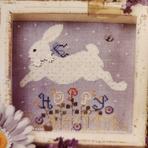 May include: Cross-stitch art of a white rabbit leaping across a lavender background with the words "HAPPY EASTER". The rabbit has a blue scarf and is surrounded by small white dots. The artwork is framed in a white, distressed wooden frame.
