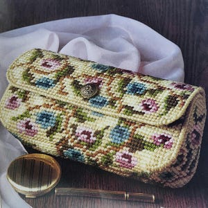 Può includere: Una pochette rettangolare beige con un motivo floreale rosa, blu e verde. La pochette ha una patta superiore arrotondata fissata con un bottone decorativo. Un compatto dorato e una penna sono in primo piano.