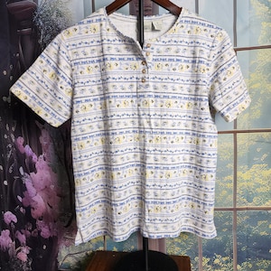 May include: A short-sleeved, button-down shirt with a floral pattern. The shirt features a cream-colored base with horizontal stripes of blue and yellow floral designs. The shirt has a round neckline and is hanging on a wooden hanger.