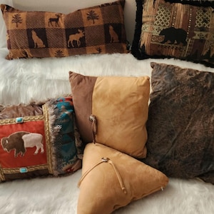 May include: A collection of decorative throw pillows in various shapes and sizes. Pillows feature animal and nature-themed designs, including bears, moose, and buffalo. Colors include brown, tan, and rust, with textured and patterned fabrics. Some pillows have decorative trim.