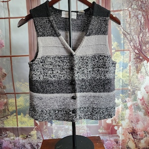 May include: A sleeveless, button-down sweater vest in shades of gray, black, and off-white. The vest features a V-neck and a striped pattern. The brand name "FIRST SOUR" is visible on the label. The vest is displayed on a wooden hanger.