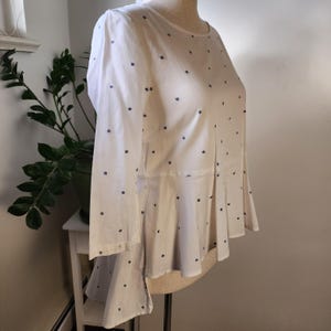 May include: A white blouse with a blue polka dot pattern. The blouse has a fitted bodice and a flared hem. The sleeves are three-quarter length.