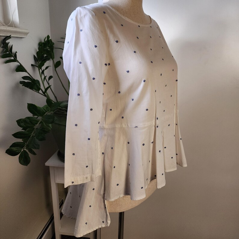 May include: A white blouse with a blue polka dot pattern. The blouse has a fitted bodice and a flared hem. The sleeves are three-quarter length.