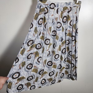 May include: A vintage midi skirt with a white background and a repeating pattern of brown circles, olive green rectangles, and blue abstract shapes. The skirt is pleated and hangs from a hanger.