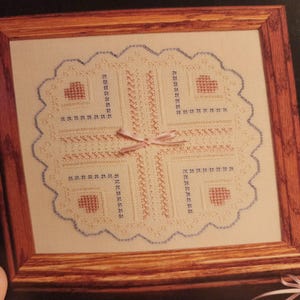 May include: A framed cross-stitch piece with a scalloped edge. The design features a central cross pattern, four corner hearts, and decorative stitching in red, blue, and cream colors. A small pink ribbon is tied at the center.