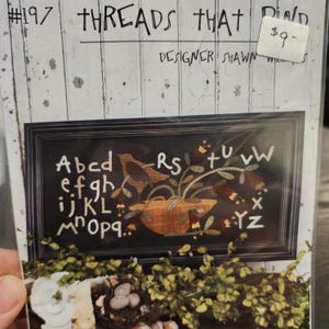 May include: A cross-stitch pattern featuring a framed design with a bird, flowers, and letters. The text "THREADS THAT" and "DESIGNER SHAWN" are visible. The price tag shows $9-.