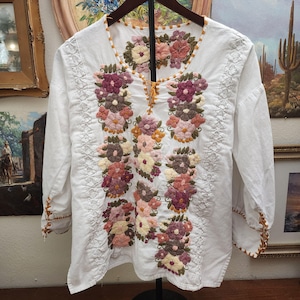 Puede incluir: Blusa blanca bordada con mangas largas. La blusa presenta bordados florales coloridos en tonos rosa, morado y marr&oacute;n. El escote y los pu&ntilde;os est&aacute;n adornados con una costura decorativa marr&oacute;n.