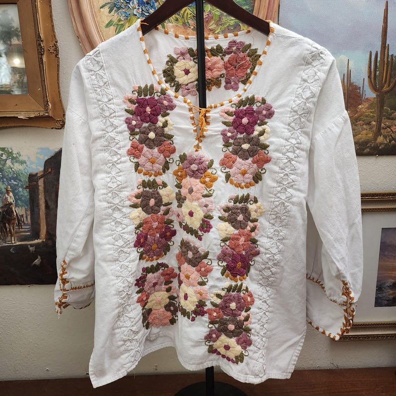 Puede incluir: Blusa blanca bordada con mangas largas. La blusa presenta bordados florales coloridos en tonos rosa, morado y marr&oacute;n. El escote y los pu&ntilde;os est&aacute;n adornados con una costura decorativa marr&oacute;n.