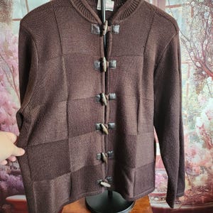 May include: A brown knit cardigan with a round neckline and long sleeves. The sweater features a series of wooden toggle closures and leather-like accents. The front of the sweater has a patchwork design with varying shades of brown.