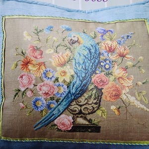 May include: Cross-stitch embroidery featuring a blue parrot perched near a vase overflowing with colorful flowers. The flowers include pink roses, orange lilies, and blue blossoms. The artwork is framed with a light blue border and green trim.