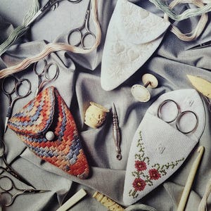 May include: A collection of sewing tools including three triangular fabric scissor cases with embroidery, multiple pairs of metal scissors, a small carved wooden spool, a silver stiletto, a bone needle case, and a crochet hook on a grey fabric.