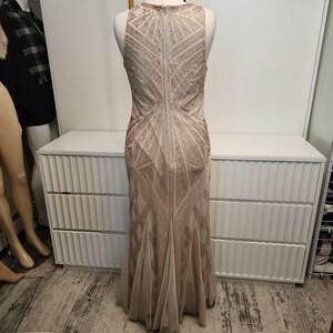 May include: A sleeveless floor-length gown in a champagne beige hue, featuring an intricate geometric pattern of gold-toned beadwork and sequins throughout the bodice and skirt, with a central vertical zipper closure along the back.