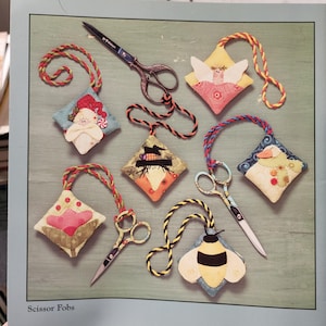 May include: A collection of decorative scissor fobs with various designs, including a Santa Claus, angel, scarecrow, duck, flower, and bee. Each fob is diamond-shaped and attached to a pair of small scissors with a braided cord. The text "Scissor Fobs" is at the bottom.