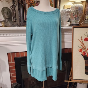 Puede incluir: Una blusa de punto de gofre de color turquesa con mangas largas, cuello ancho y aberturas laterales. La camisa tiene un corte holgado y un dobladillo ligeramente con volantes. La tela tiene un aspecto texturizado.