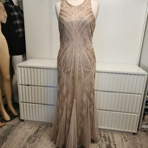 May include: A floor-length sleeveless gown in a light beige hue, featuring intricate gold beadwork arranged in geometric patterns across the bodice and skirt. The dress has a high neckline and a flared hemline made of sheer, layered fabric.