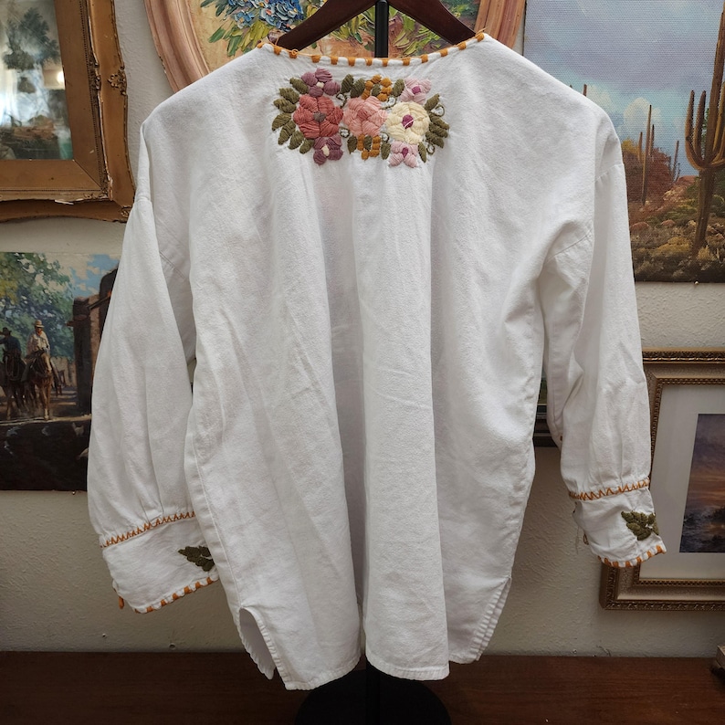 Puede incluir: Blusa blanca de manga larga con bordado floral en rosa, melocot&oacute;n y verde. El bordado est&aacute; centrado en la parte superior de la espalda y alrededor del escote. Los pu&ntilde;os y el escote est&aacute;n adornados con costuras naranjas y verdes.