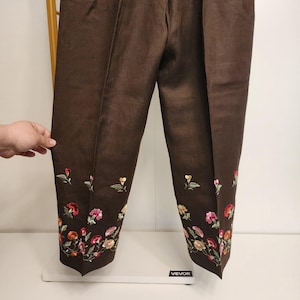 May include: Brown pants with floral embroidery along the hem. The embroidery features a variety of colorful flowers, including red, pink, and yellow. The pants are made of a textured fabric and have a classic, tailored fit.