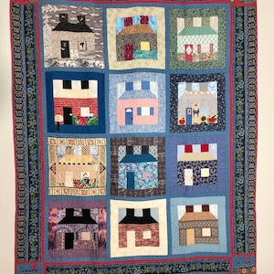 May include: A quilt with a blue background and a red border. The quilt is made up of 12 squares, each with a different design of a house. The houses are made of different fabrics and colors, and some have windows and doors.