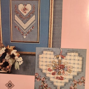 May include: A framed cross-stitch design featuring a heart and floral elements. The design is set against a blue background with a gold-colored frame. A smaller cross-stitch heart design is also visible.