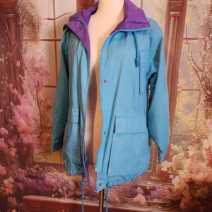 May include: A light blue jacket with a purple hood and lining. The jacket has a zipper closure, two front pockets, and drawstrings at the hood and waist. The jacket is displayed on a mannequin.