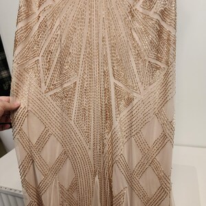 May include: A floor-length dress in a light beige hue featuring an intricate geometric pattern created with metallic gold beads. The beadwork is arranged in symmetrical lines and angular shapes that extend vertically across the entire fabric surface.