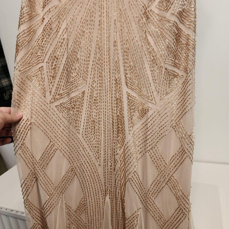 May include: A floor-length dress in a light beige hue featuring an intricate geometric pattern created with metallic gold beads. The beadwork is arranged in symmetrical lines and angular shapes that extend vertically across the entire fabric surface.