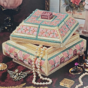 May include: A square plastic canvas box with a removable lid, featuring a geometric pattern in cream, pink, and teal yarn. A string of white faux pearls hangs over the front edge of the box, which is surrounded by various small vintage jewellery items.