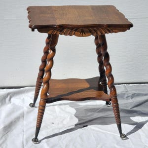 May include: A vintage wooden side table with a dark brown finish. The table features a square top with a scalloped edge, ornate carved details, and spiral-turned legs. It has a lower shelf and small caster wheels on the feet.