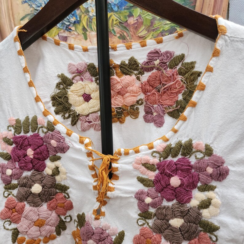 Puede incluir: Top blanco bordado con dise&ntilde;os florales coloridos. El bordado presenta tonos de rosa, morado, marr&oacute;n y verde. El cuello y los bordes est&aacute;n adornados con costuras naranjas. La parte superior est&aacute; colgada en una percha de madera.