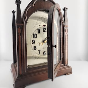 May include: A wooden mantel clock with an arched top and a glass door. The clock face is silver with black numerals and gold hands. The clock is dark brown with decorative trim and finials. The door is open, revealing the clock face.