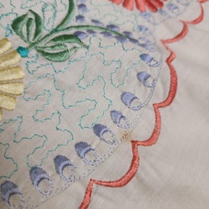 May include: Close-up of a cream-colored textile with intricate embroidery. The design features floral and geometric patterns in shades of yellow, green, blue, and coral. The edges are scalloped with coral stitching, adding a decorative touch to the fabric.