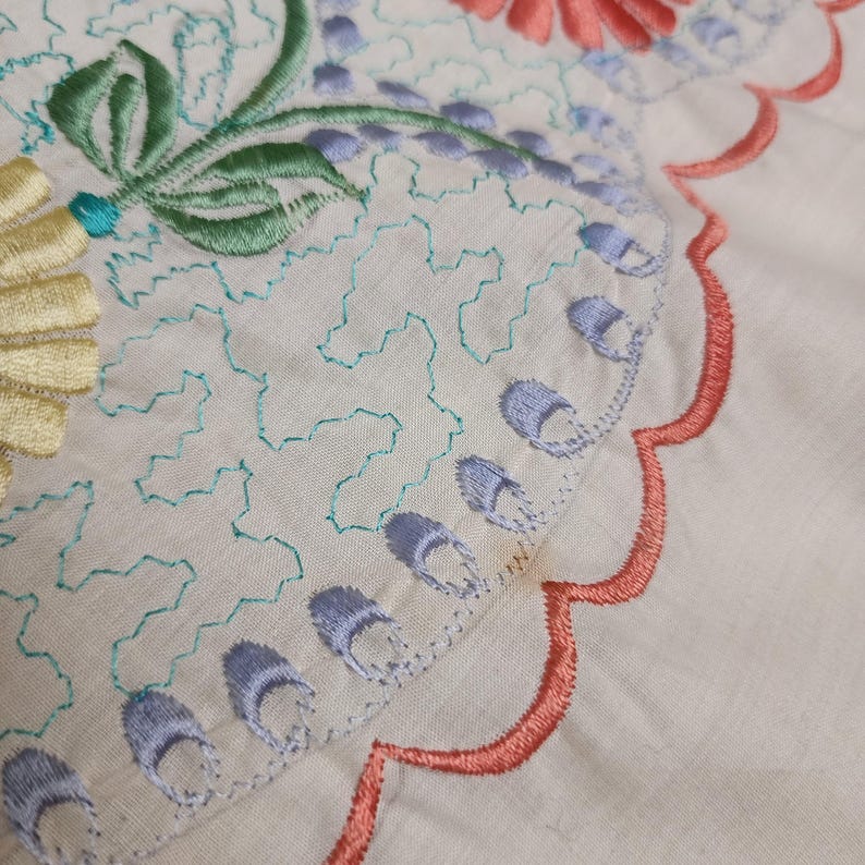 May include: Close-up of a cream-colored textile with intricate embroidery. The design features floral and geometric patterns in shades of yellow, green, blue, and coral. The edges are scalloped with coral stitching, adding a decorative touch to the fabric.
