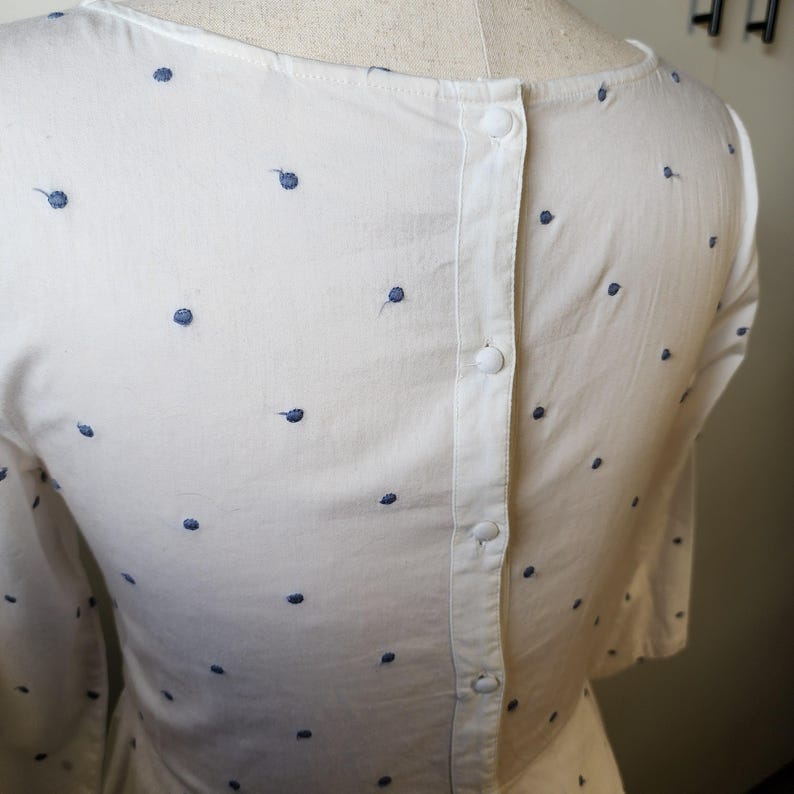 May include: White button-down shirt with a polka dot pattern. The shirt has a button closure down the back.