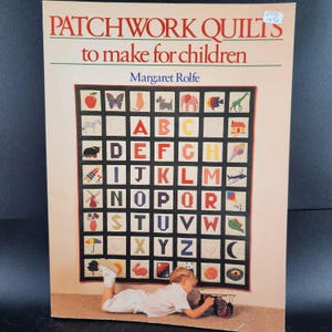 May include: A book cover titled "PATCHWORK QUILTS to make for children" by Margaret Rolfe. The cover features a colorful patchwork quilt design with the alphabet and various images. A child is lying on the floor near the book.