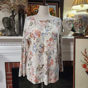 May include: A long-sleeved, cream-colored top with a floral pattern in shades of pink, blue, and purple. The top has a round neckline and is displayed on a mannequin. The floral print is spread across the entire top.