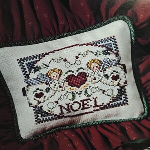 Puede incluir: Un cojín rectangular de punto de cruz con dos figuras de cabello rubio que sostienen un gran corazón rojo oscuro. El diseño incluye enredaderas verdes, detalles azules en las esquinas y la palabra NOEL bordada en hilo rojo oscuro sobre tela blanca.