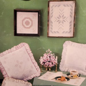 May include: A display of vintage-style home decor items. Two framed white lace doilies hang on a green wall. Two pink lace-trimmed pillows and a third pillow are on a table with a floral arrangement, tea, and cookies.