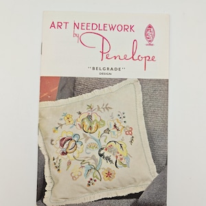 May include: A rectangular instructional booklet titled Art Needlework by Penelope Belgrade Design. The cover features a cream-coloured square pillow with a fringed edge, decorated with a colourful floral embroidery pattern in yellow, blue, and green.