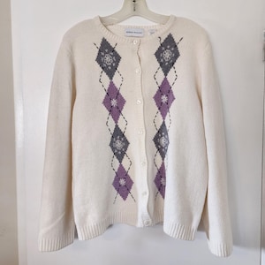 May include: A white cardigan sweater with a diamond pattern in purple and gray. The sweater has a button closure and long sleeves.