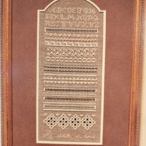 May include: A framed sampler with various embroidered stitch patterns in white thread on a tan background. The top features the alphabet, and the bottom has the text "A stitch in time". The frame is a light brown color.